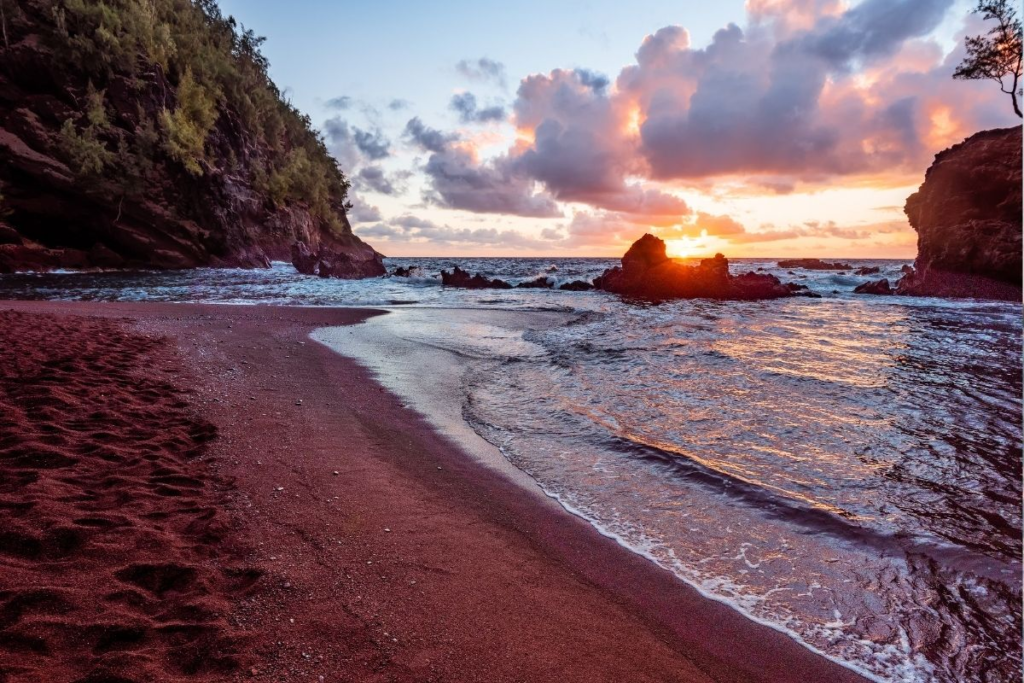 Sunset over a red sand beach with gentle waves, rocky cliffs, and lush greenery on the left. The sun is partially hidden behind rocks on the horizon, casting a warm glow on the clouds and water.