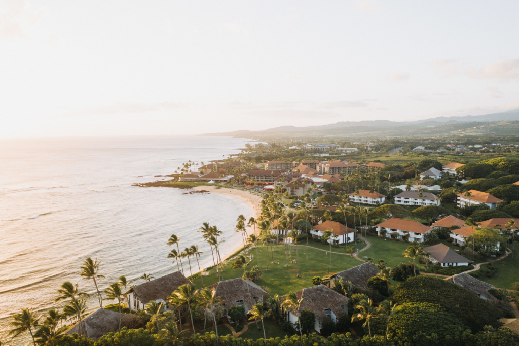 Aerial view of a tropical coastline with sandy beach, palm trees, and resort buildings; lush greenery surrounds the area, and gentle ocean waves approach the shore under a soft, golden sunset sky.
