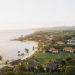 Aerial view of a tropical coastline with sandy beach, palm trees, and resort buildings; lush greenery surrounds the area, and gentle ocean waves approach the shore under a soft, golden sunset sky.