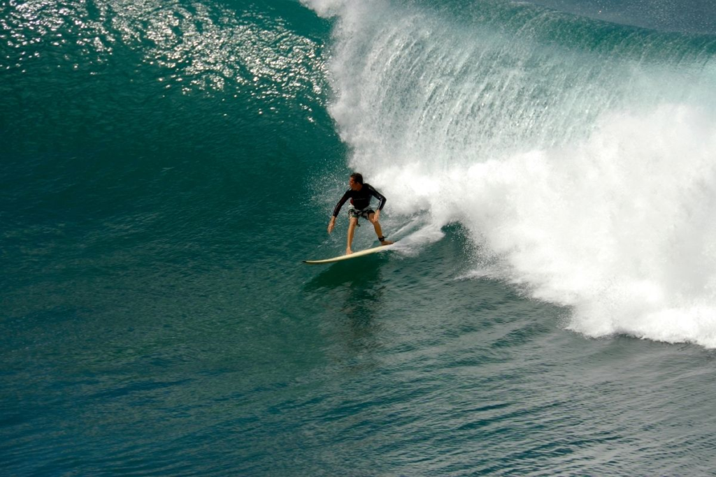 A surfer in a black wetsuit rides inside the barrel of a large, turquoise wave near the romantic hotels in Maui, surrounded by spray and sunlight reflecting off the water.