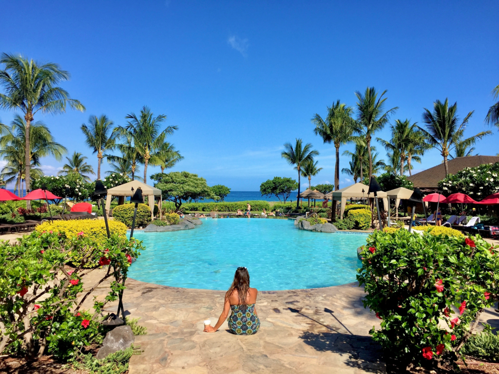 A woman sits at the edge of a tropical resort pool surrounded by lush greenery and palm trees, with umbrellas and lounge chairs nearby, and the ocean visible in the distance under a clear blue sky.