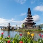 A lakeside Balinese temple with tiered roofs sits on a small island, surrounded by water and lush green hills. Bright red and yellow flowers bloom in the foreground under a blue sky with scattered clouds.