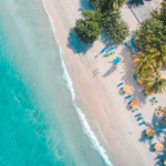 Aerial view of a tropical beach with turquoise water, white sand, sun umbrellas, lounge chairs, green palm trees, and a red-roofed building surrounded by lush vegetation.